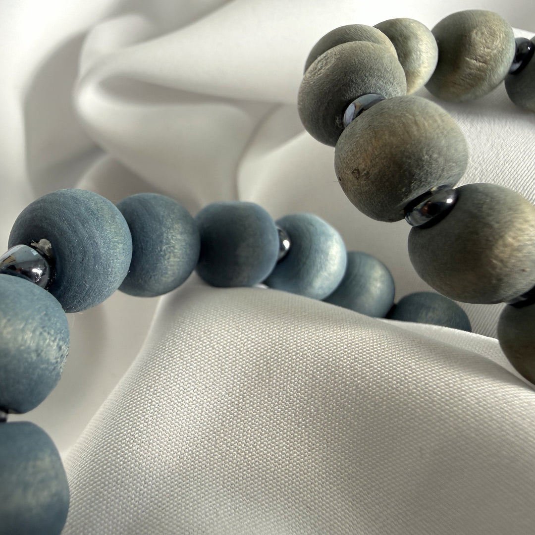 Close-up of two handcrafted bracelets laid side by side showing blue and grey wood beads with silver-coloured seed bead accents.