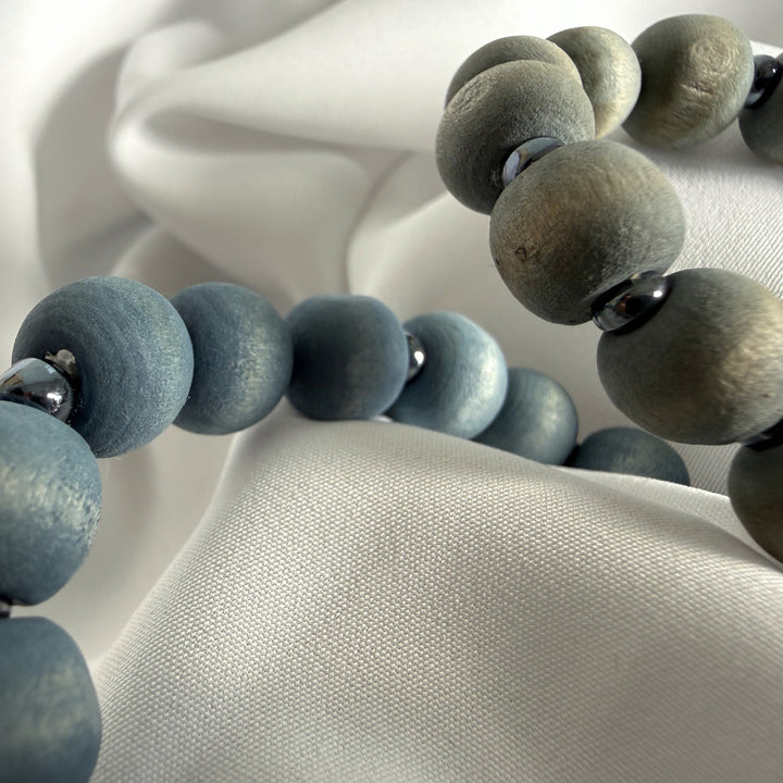 Close-up of two handcrafted bracelets laid side by side showing blue and grey wood beads with silver-coloured seed bead accents.