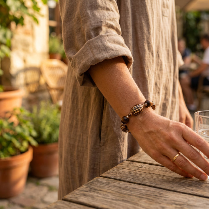 Handcrafted beaded bracelet with ceramic, wood, glass, and acrylic beads worn on a woman’s wrist outdoors while drinking water in a calm setting with plants and people sitting nearby.
