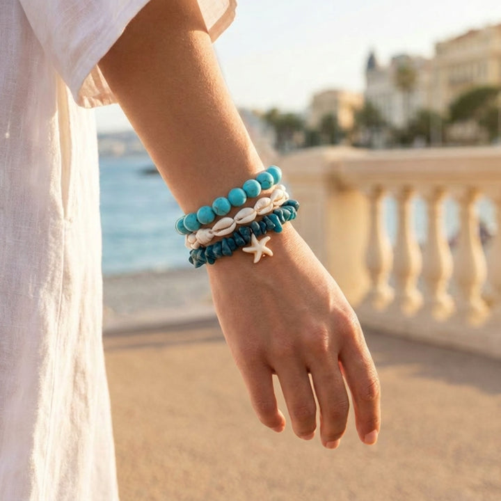 Set of three handcrafted coastal-inspired bracelets with turquoise howlite, cowrie shells, and blue chip beads worn on a wrist while walking along the beach promenade in Nice.