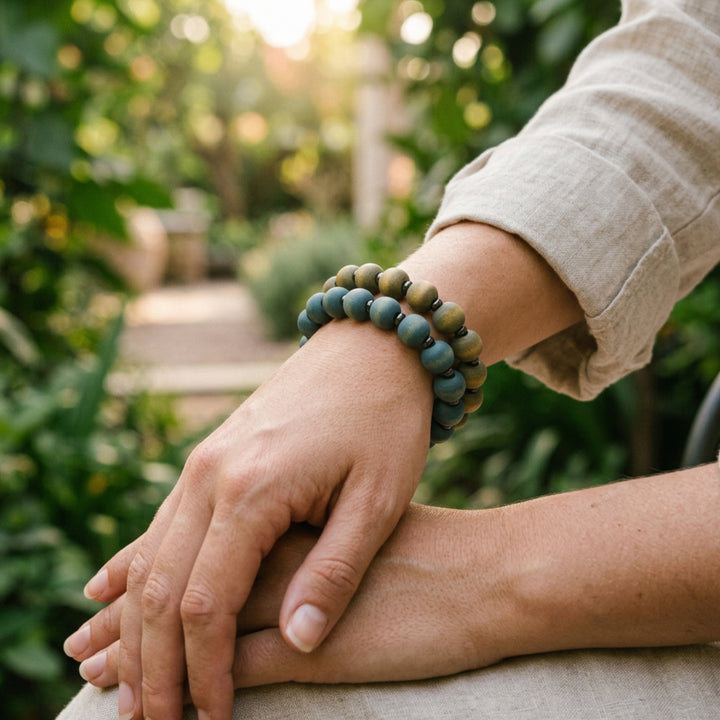 Set of two handcrafted beaded bracelets in cool blue and grey tones worn on a woman’s wrist in an outdoor garden setting.