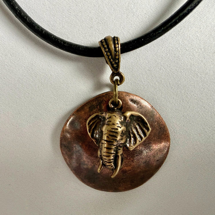 Handcrafted necklace featuring a copper disc pendant layered with an antique brass elephant charm on black leather cord.