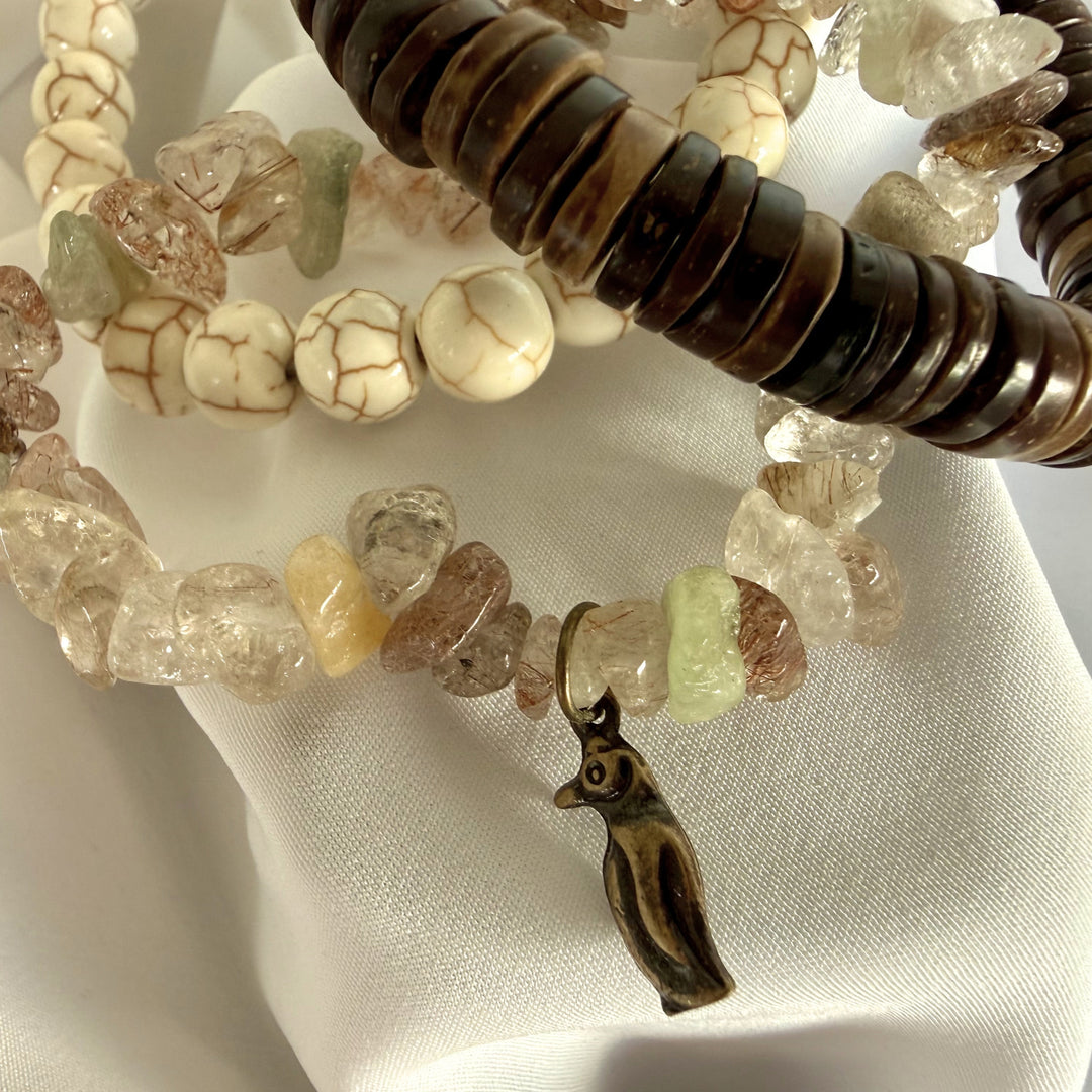 Close-up of layered howlite, coconut wood, and mother of pearl bracelets with a small penguin charm.