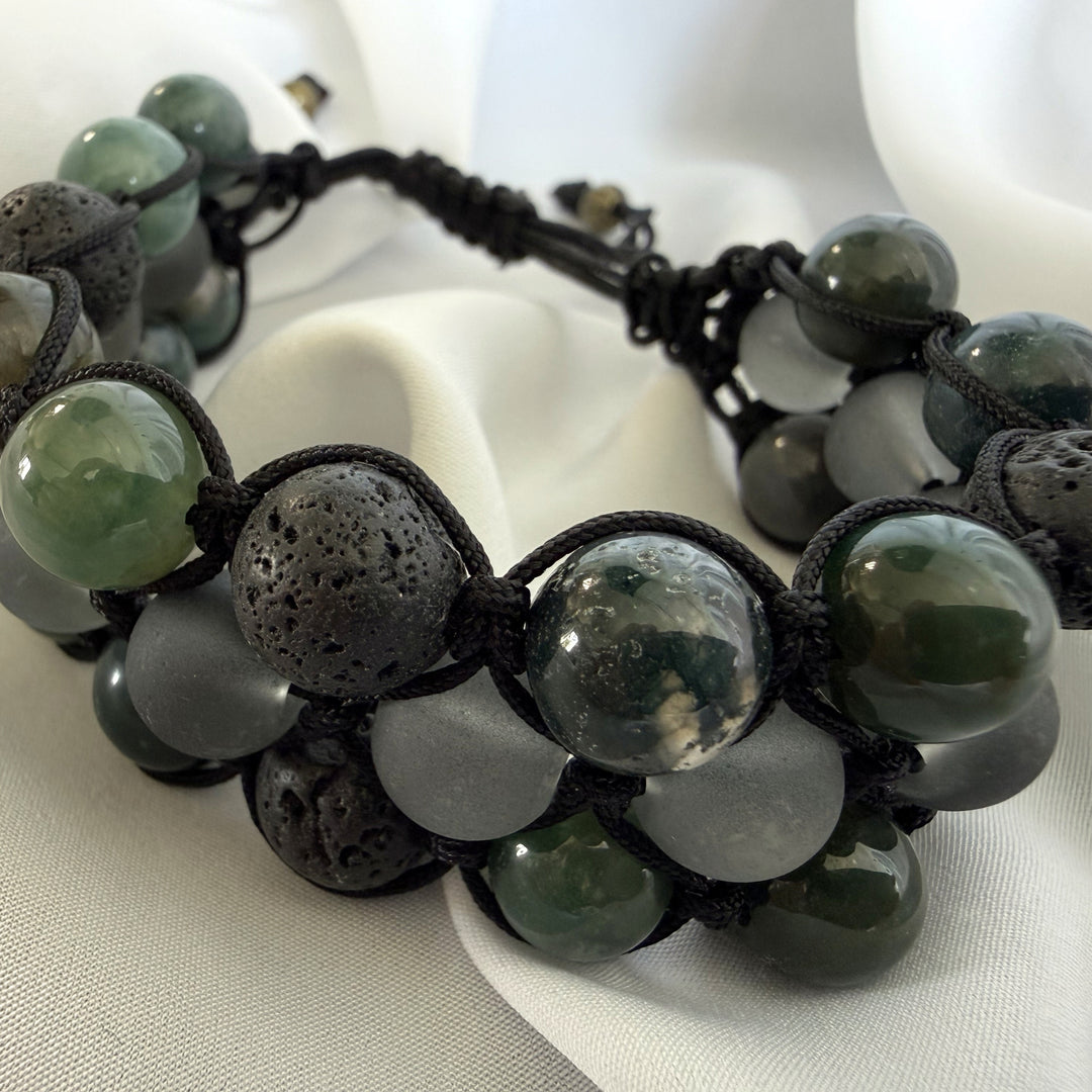 Top angled full view of a handcrafted moss agate macramé bracelet with green stones, lava beads, and black accents.