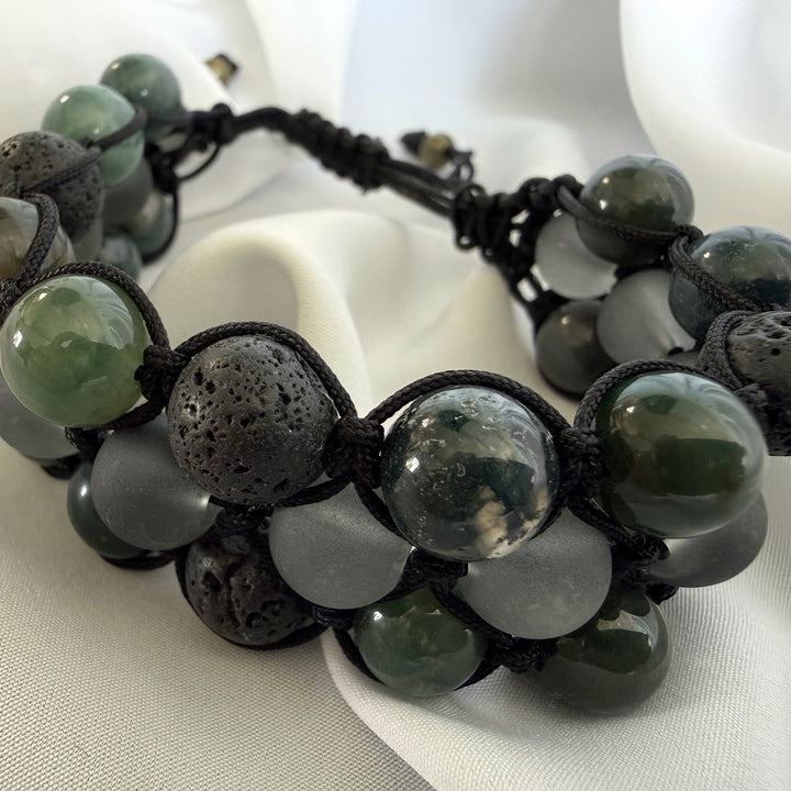 Top angled full view of a handcrafted moss agate macramé bracelet with green stones, lava beads, and black accents.