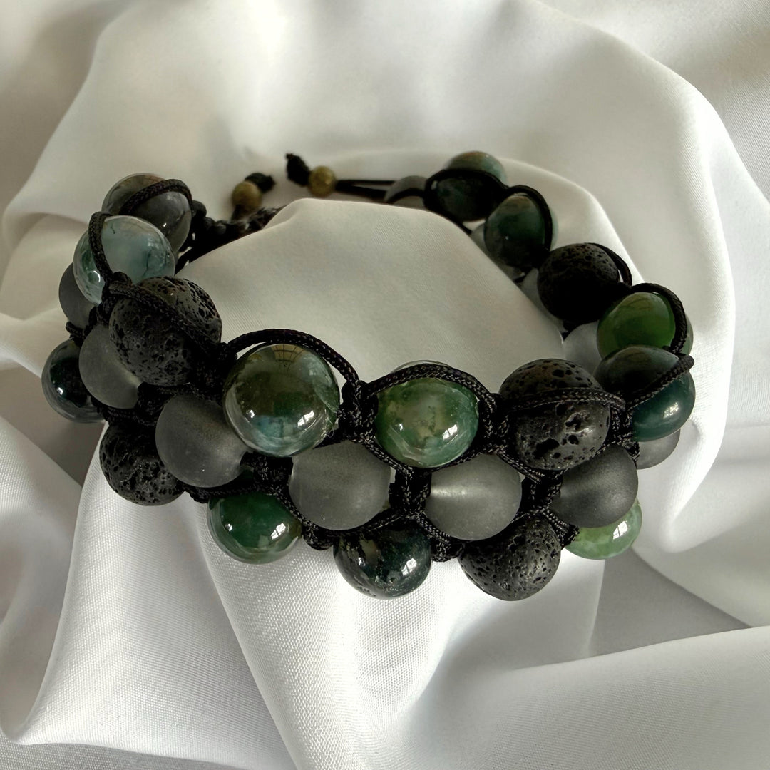 Handcrafted moss agate macramé bracelet with lava beads and black glass beads on white silk fabric.