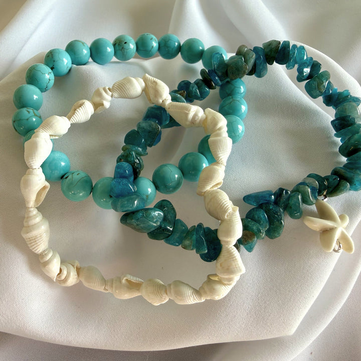 Full view of three handcrafted bracelets layered together with turquoise howlite beads, cowrie shells, and blue chip beads.