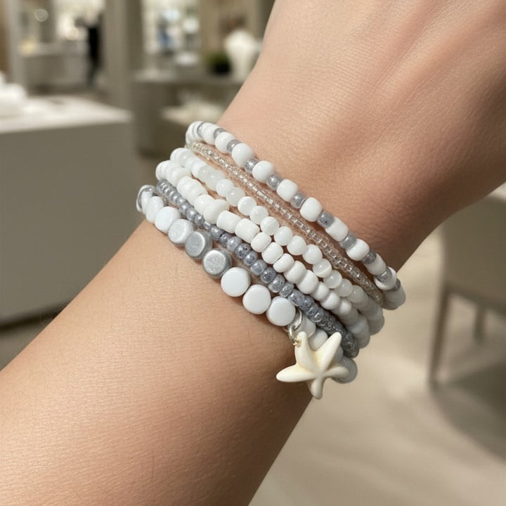 Set of six handcrafted stackable bracelets with silver-tone and white beads worn on a woman’s wrist with a white seastar charm detail.