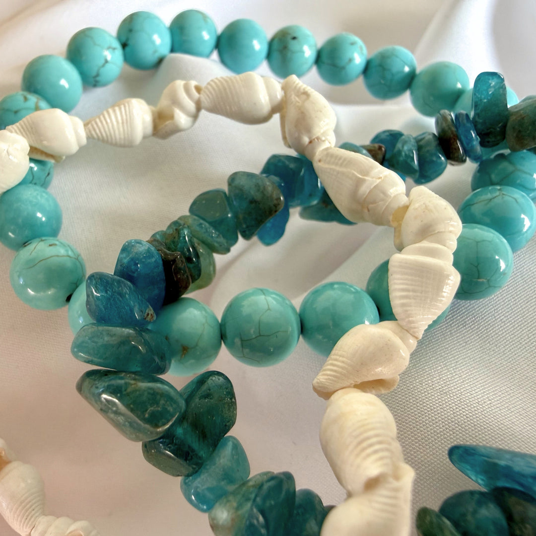 Top close-up view of layered turquoise howlite, cowrie shell, and blue chip bead bracelets spread out together.