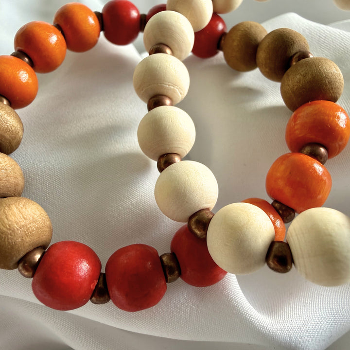 Close-up of two layered handcrafted bracelets in red, orange, gold, brown, and ivory tones made with wood and seed beads.
